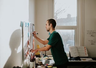 A man leaning over his desk and writing on a whiteboard in a sunny home office room. Learn "how long does it take to form a habit?" in this article.