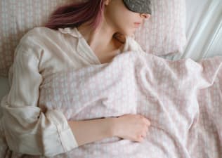 A woman with pink hair and an eye mask sleeping soundly in bed at the best temperature for sleep.