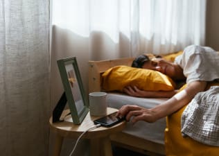 In this image for an article about how to fix your sleep schedule, a sleepy woman is lying in bed and reaching out to turn off the morning alarm on her phone. 