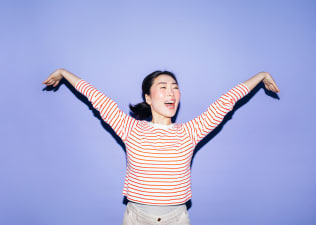 A woman smiling and spreading out her arms energetically. She's wearing a red and white striped shirt and standing against a lavender background. Learn how to get more energy in this article.