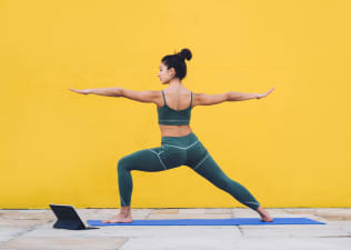 Side view of focused calm female in sportive outfit concentrating in warrior pose on yoga mat with yellow background behind