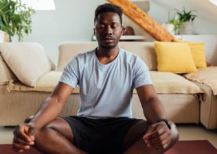 A man peacefully meditating in the morning. He's sitting on a yoga mat in front of his couch in a sunny living room.