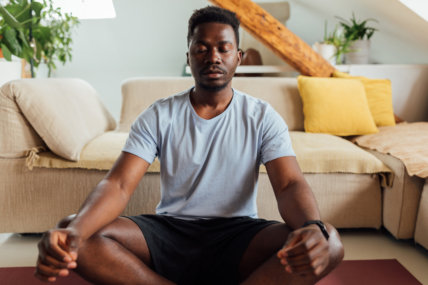 A man peacefully meditating in the morning. He's sitting on a yoga mat in front of his couch in a sunny living room.