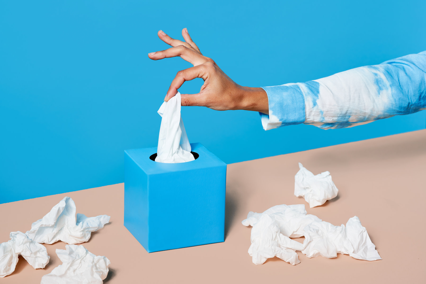In this image for an article about whether or not you should exercise when you're sick, a hand in a blue and white tie-dye jacket is plucking a tissue from a tissue box. There are used tissues lying around the tissue box on a cream table.