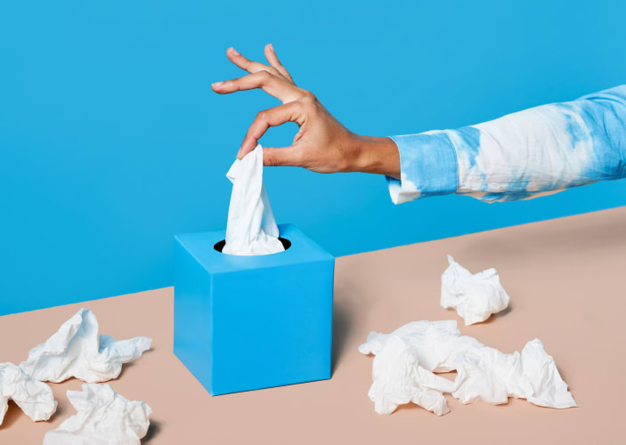 In this image for an article about whether or not you should exercise when you're sick, a hand in a blue and white tie-dye jacket is plucking a tissue from a tissue box. There are used tissues lying around the tissue box on a cream table.