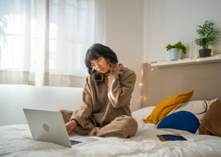 A woman experiencing tech neck. She is rubbing her neck while sitting in bed and looking down at her laptop.