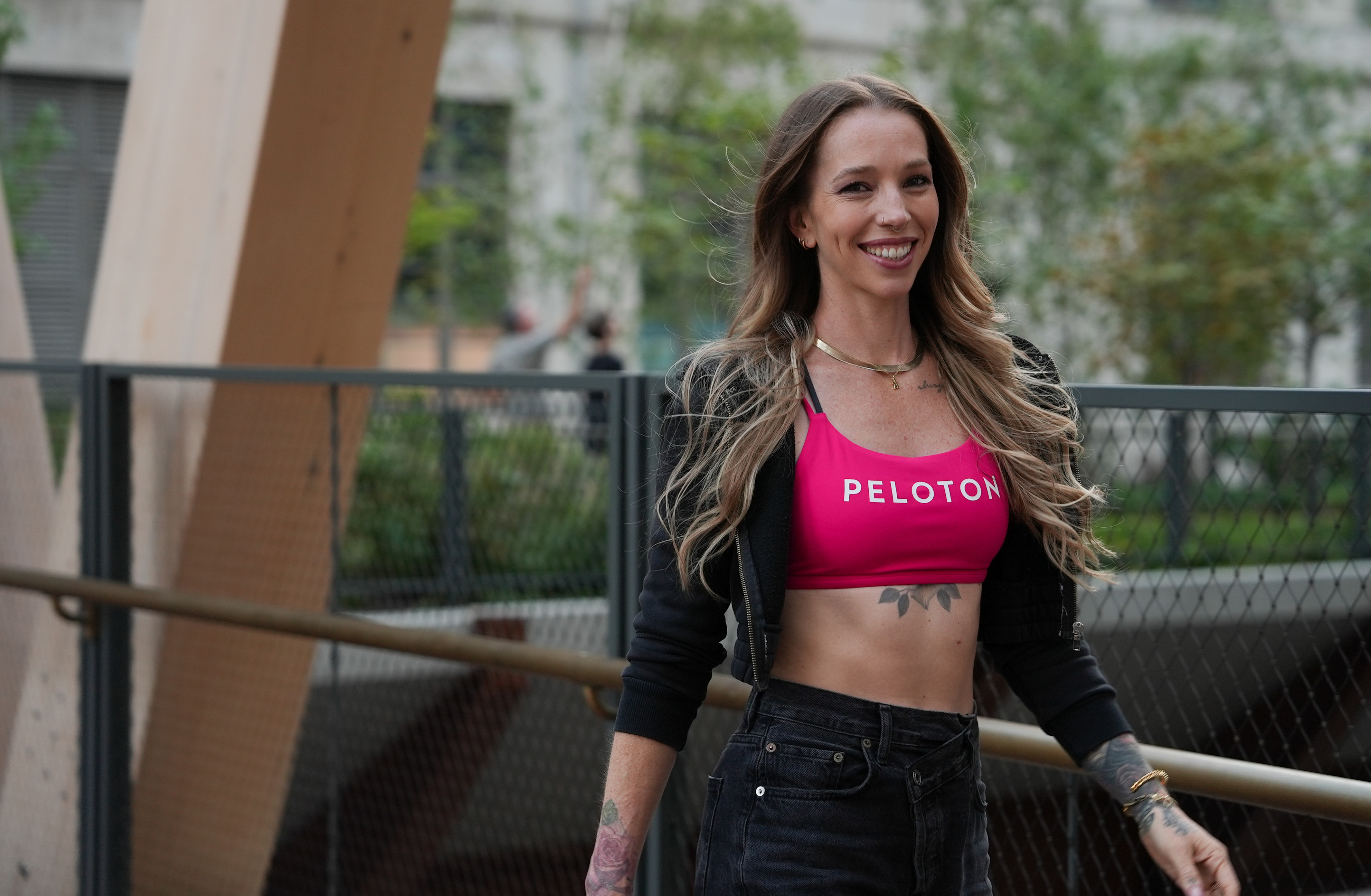 Peloton instructor Kirra Michel going on a walking meditation outdoors. She's smiling and wearing a pink Peloton sports bra, a black zip-up hoodie, and black jeans.