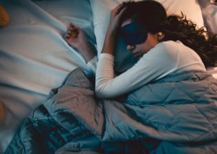 A woman sleeping in bed peacefully while wearing an eye mask and snoozing under a weighted blanket. Weighted blankets offer many benefits.