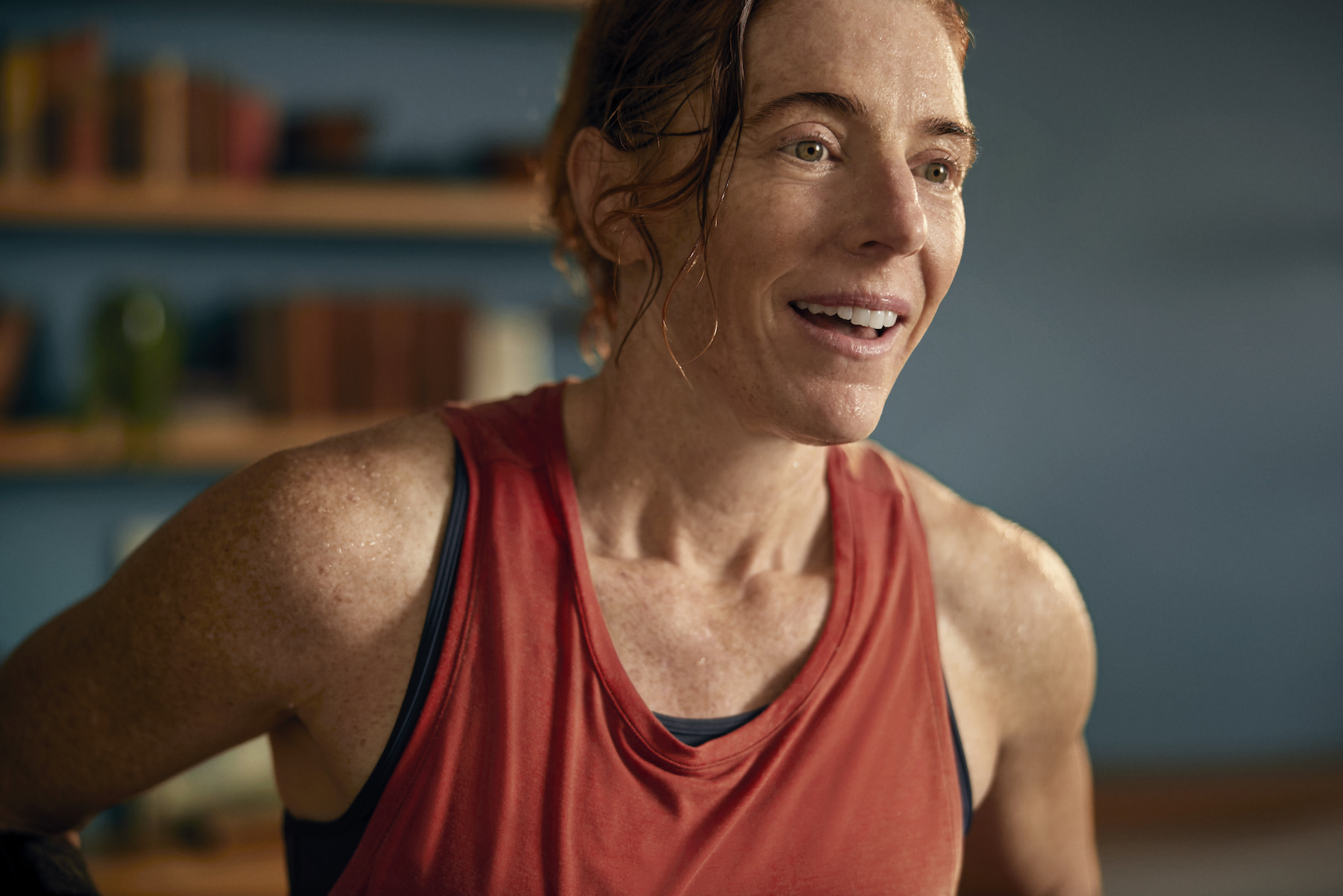 A woman smiling after finishing a home workout. She is sweaty and wearing a red tank top.